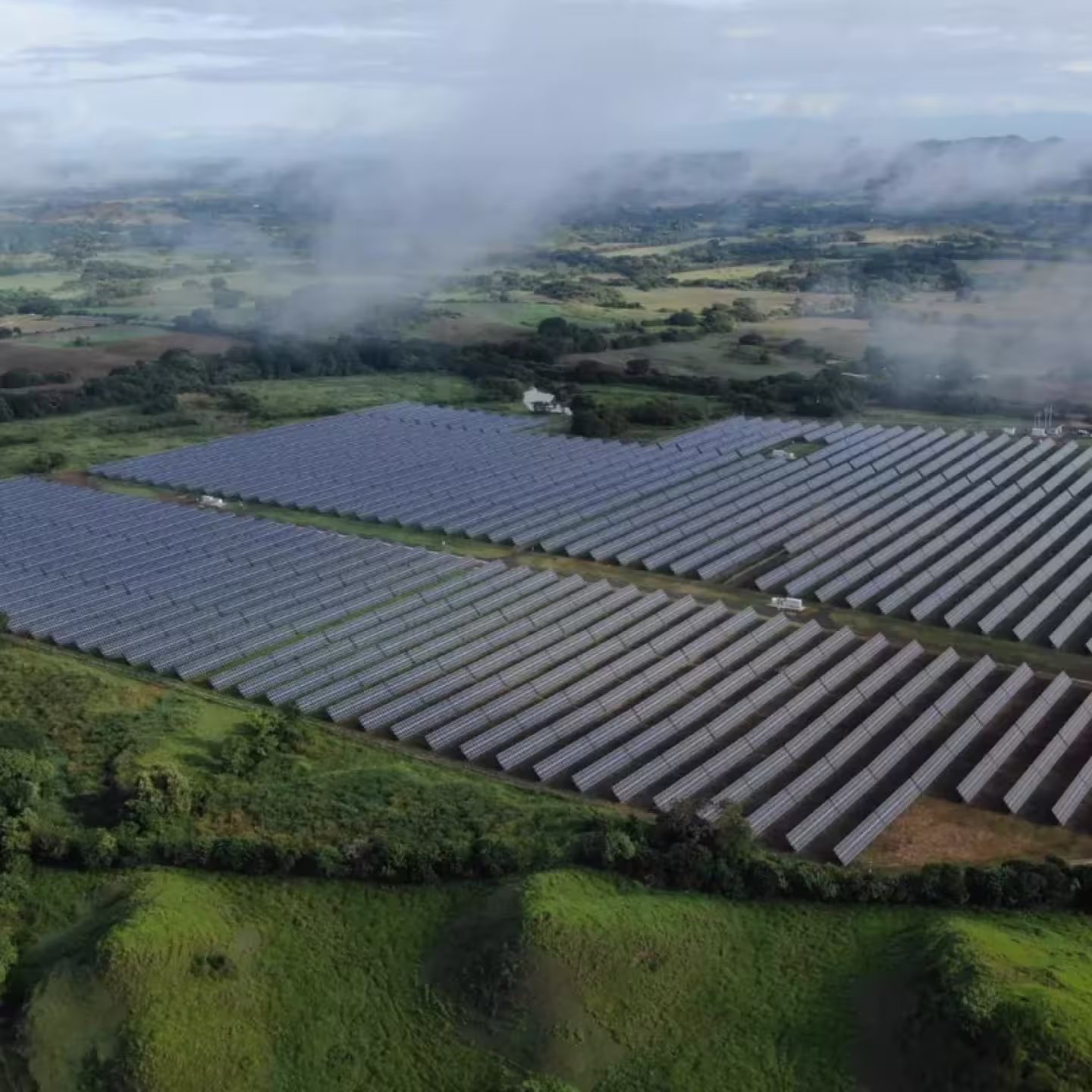 Aerial view of a large solar panel farm surrounded by green fields and a misty landscape.